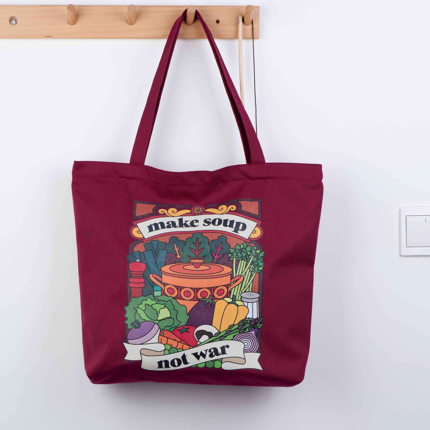 Burgundy tote bag made from canvas with vintage retro style design on the front saying Make Soup Not War, with an illustration of soup ingredients and an old style pot. It has a zip across the top and a pocket inside. Could be a shopping bag or a handbag. 