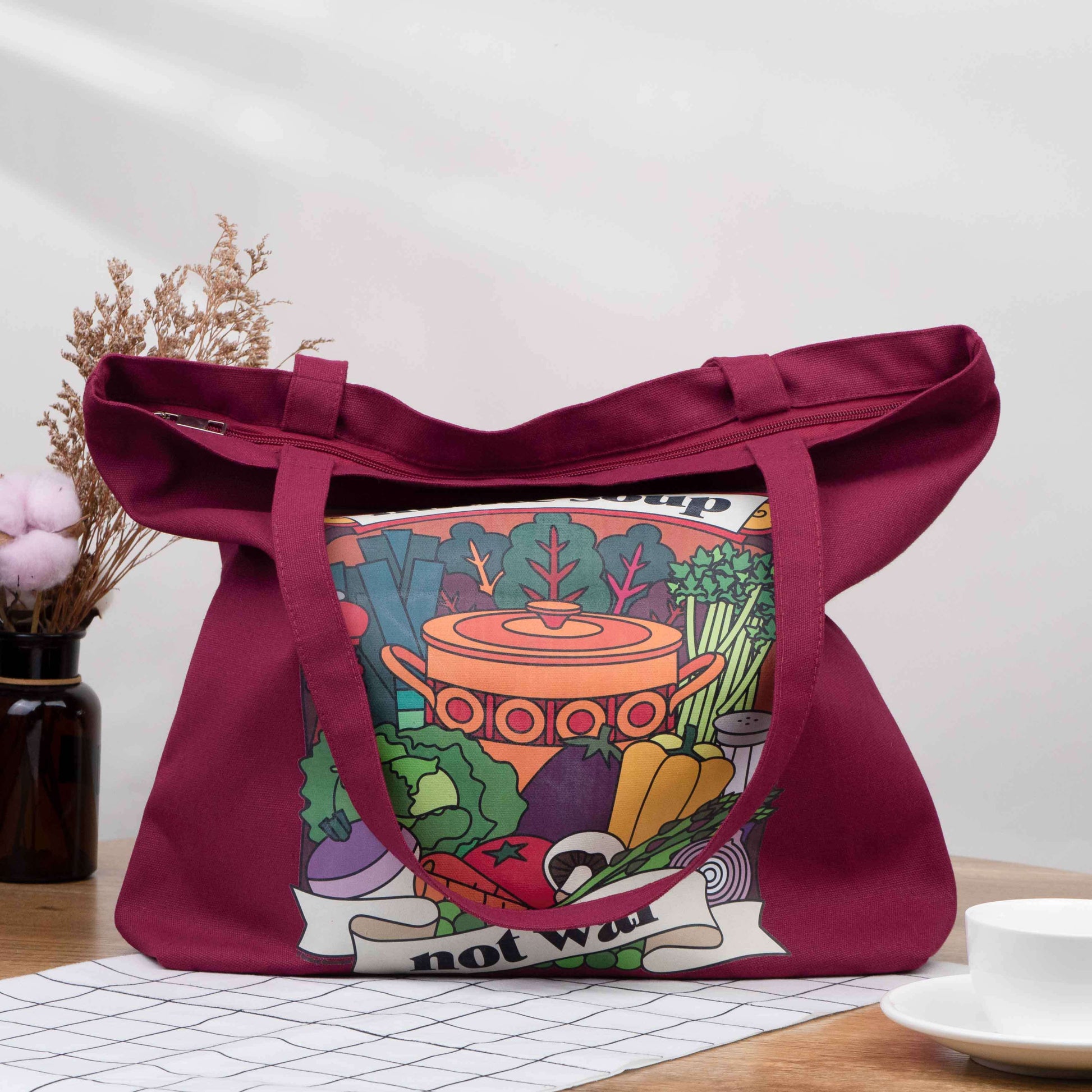 Burgundy tote bag made from canvas with vintage retro style design on the front saying Make Soup Not War, with an illustration of soup ingredients and an old style pot. It has a zip across the top and a pocket inside. Could be a shopping bag or a handbag. 