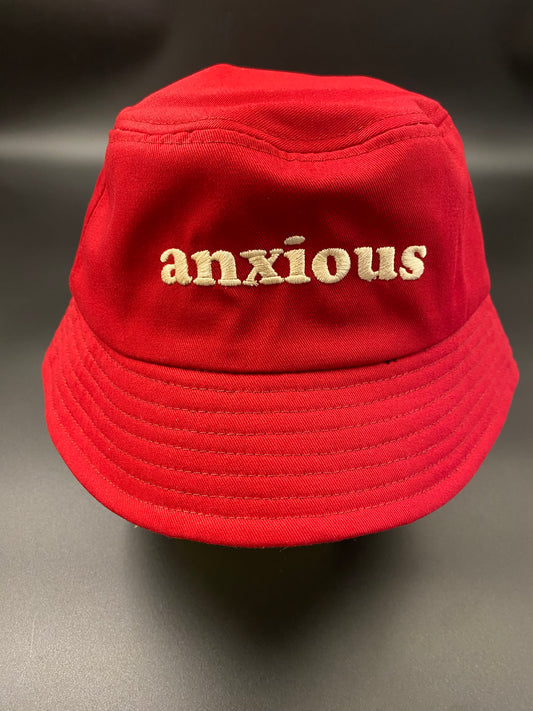 Bucket of Anxiety: Cherry red heavy cotton bucket hat with neutral coloured embroidery across the front reading ANXIOUS. Has an internal sweatband and lines sewed across the brim. 